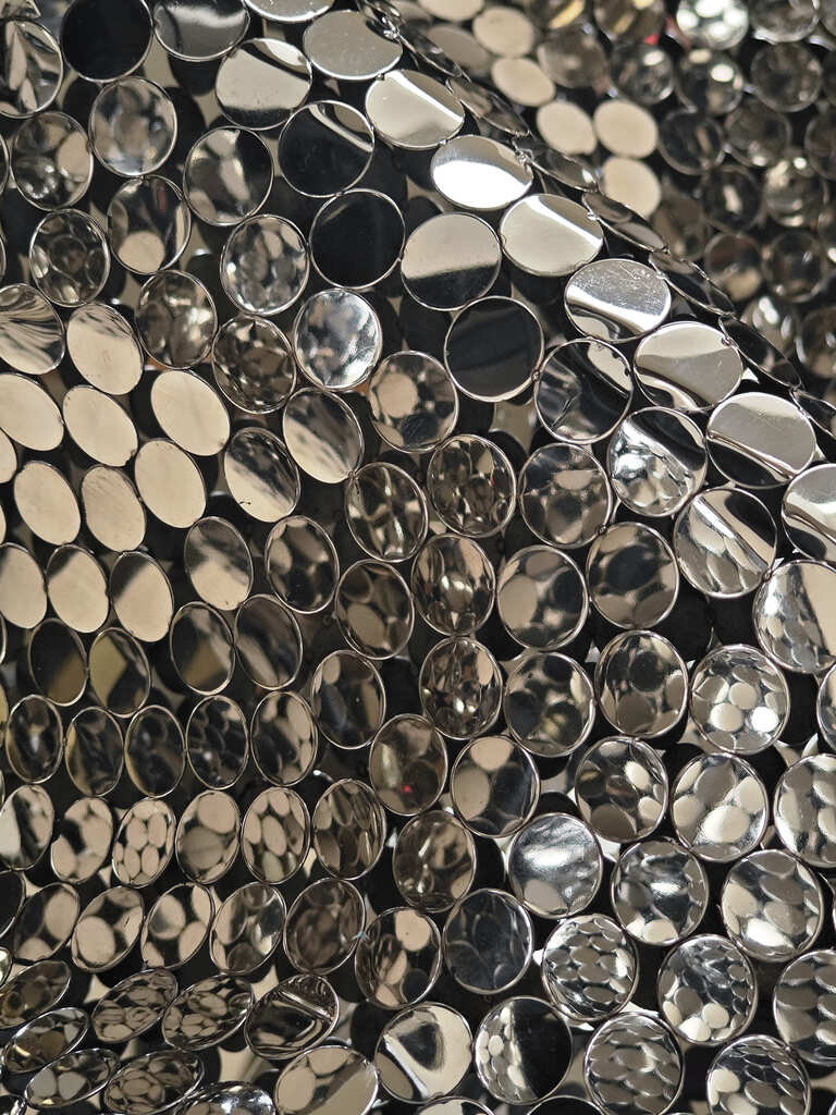 A close-up of a fabric covered in shiny, round silver sequins, creating a reflective, metallic pattern with light and shadow.